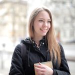 A young woman smiles while holding a book, captured in a city street setting.