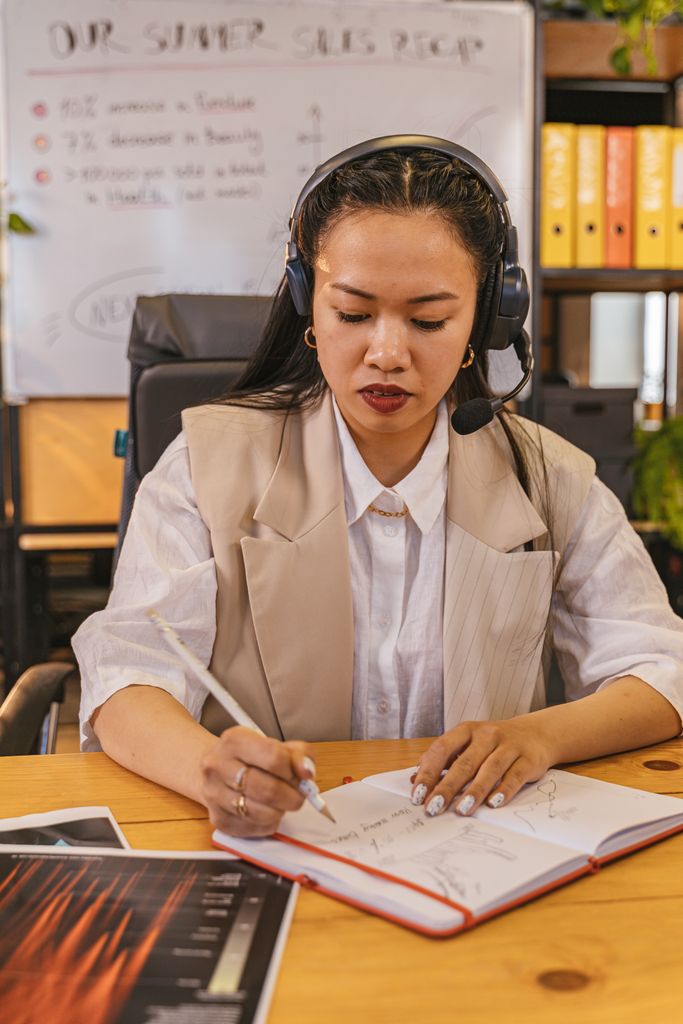 Asian woman in headset focuses on work, taking notes in office setting.